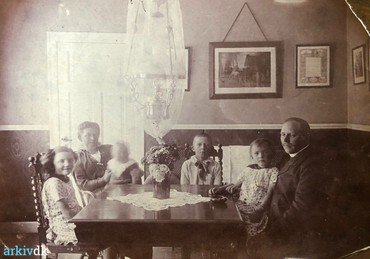 1917 - Familien Andersen i spisestuen en sommerdag (petroleumslampen fra loftet er pakket ind). Fra venstre Rigmor, Karen med Ernst på skødet, Helge og Anders med Dagny på skødet. – Billedet på væggen er en reproduktion af Otto Baches ”Et kobbel heste udenfor Lindenborg kro (1881).