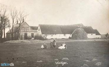 1917 – Det ældste billede af gården, set fra nord. Det grundmurede stuehus er bygget af den tidligere ejer (Jens Peter Larsen 1890-1906). Foran nordlængen er det korn, som der ikke var plads til i laden, sat ”i hæs”. – Forrest i billedet Karen Andersen med Ernst (f. 2.10. 1916) på skødet, dernæst Dagny (f. 1913) og den ældste, Rigmor (1909). Til højre tjenestepigen (navn ukendt).